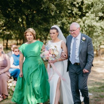 Bride in a white wedding dress walking down the ceremony aisle accompanied by both of her parents.