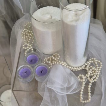 Tall white cylinder candles in glass vases arranged on a floor display with soft white fabric.